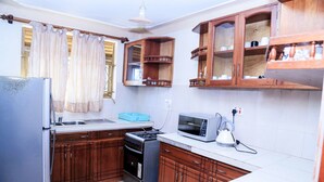 A compact kitchen with wooden cabinets, a refrigerator, a stove, a microwave, and a kettle. - Floben Apartments (Kampala)