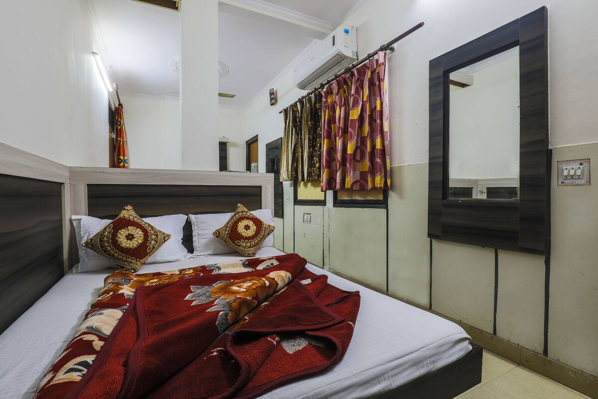 A bed with a patterned blanket, a headboard, two decorative pillows, a mirror, and a curtain.