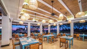 Uma sala de jantar ampla, com várias mesas e cadeiras, grandes janelas com vista para o exterior e luminárias de vime suspensas.