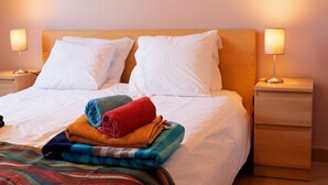 Een net opgemaakt bed met witte kussens en een stapel kleurrijke handdoeken. Aan beide zijden van het bed staan nachtkastjes met lampen.