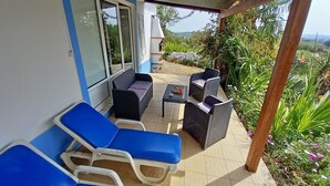 Een overdekt terras met blauwe loungestoelen en rieten meubels, omgeven door weelderige begroeiing en uitzicht op het landschap.