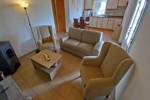 A living room with a sofa, armchairs, a coffee table, and a TV. There is a kitchen in the background with cabinets and a window.