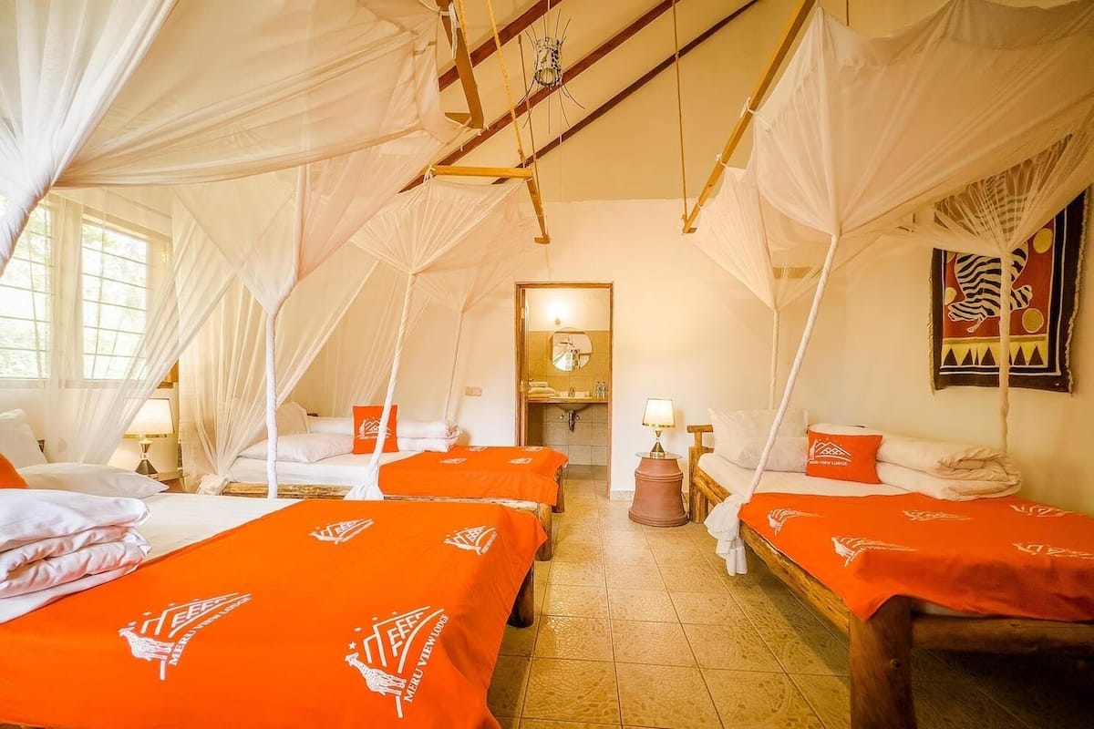 A room with four beds, each with a canopy and orange bedspreads. There is a wooden bench and a small table with a lamp. The room has a tiled floor and a large window on one side.