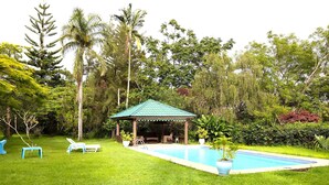 Un mirador con techo verde, una piscina y una zona ajardinada con sillones de descanso.