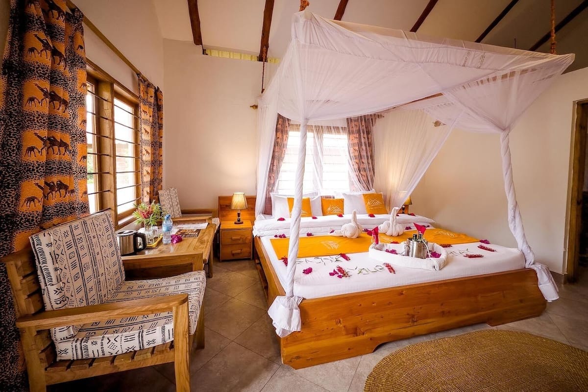 A room with a wooden bed, a wooden chair, a small table with a lamp, and a window with curtains.