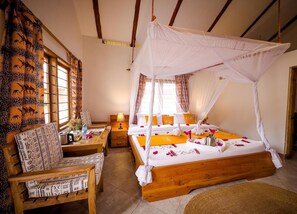 A room with a wooden bed, a wooden chair, a small table with a lamp, and a window with curtains.