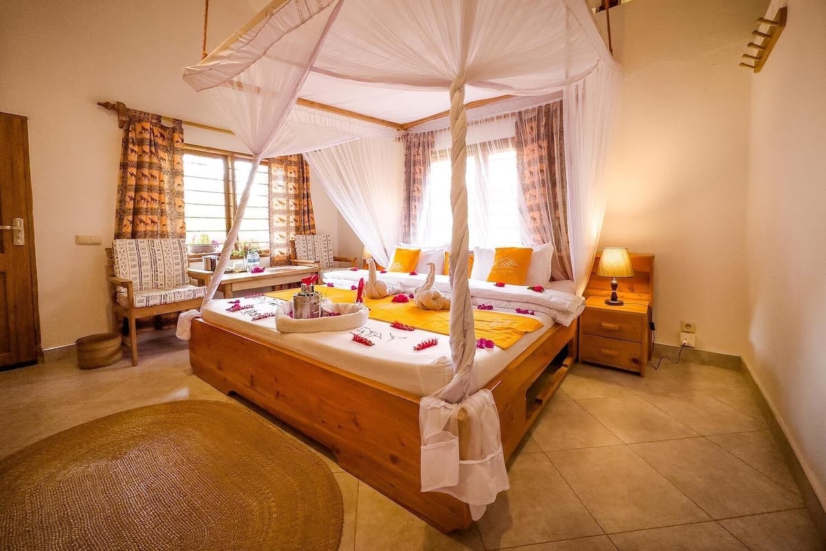 A room with a four-poster bed, a wooden bedside table, a nightstand, and a large window with curtains.