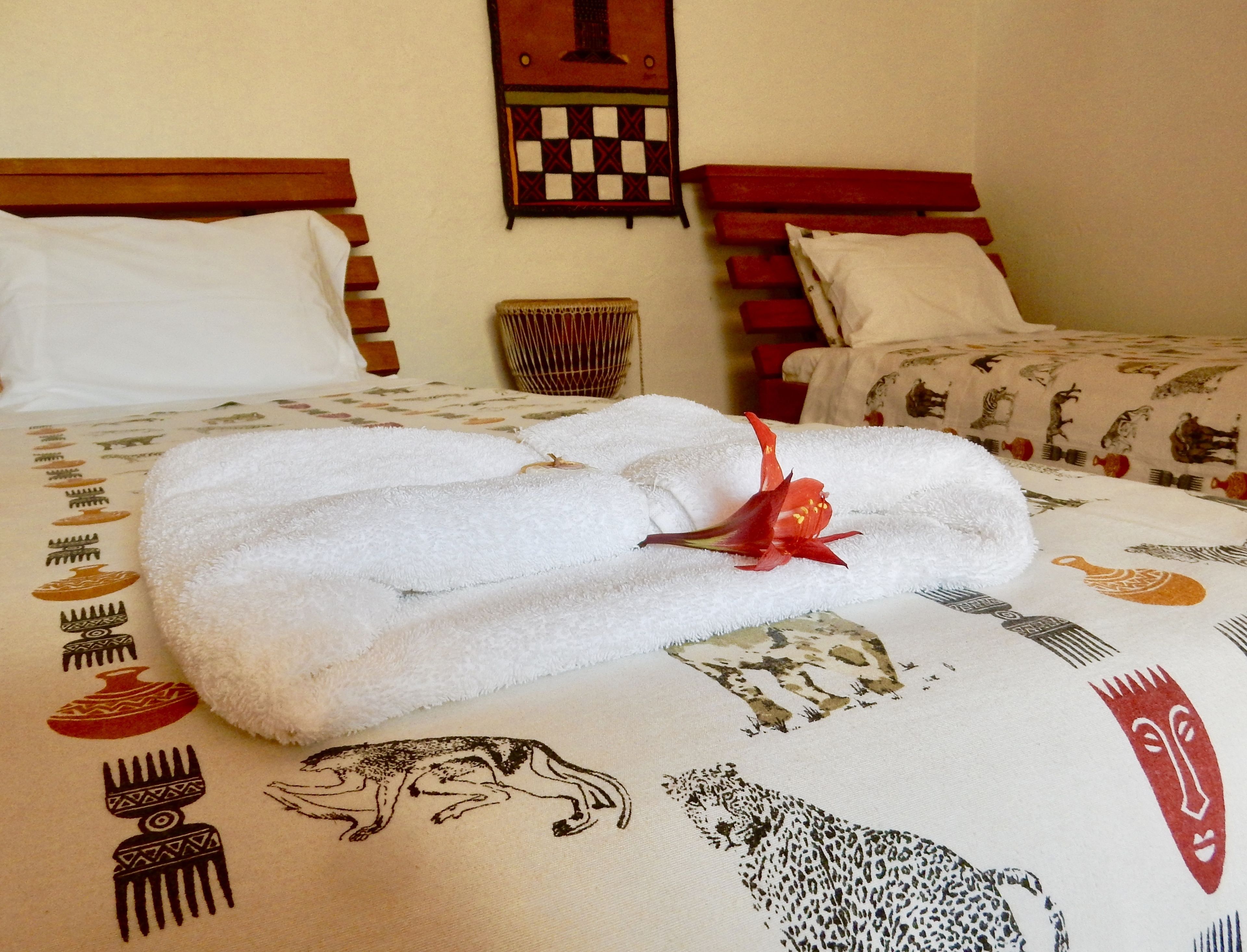 A neatly made bed with a white towel and a red flower, a wooden headboard, and a wall with a patterned tapestry.