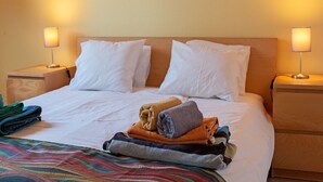Una cama bien hecha con sábanas blancas, un colorido edredón a rayas y toallas dobladas en varios colores. Hay dos lámparas de noche sobre mesitas de madera.