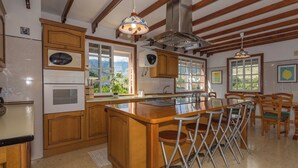 A kitchen with wooden cabinets, a central island with stools, a built-in oven, and a large window offering a view of the outdoors. - GC0181 Holiday cottage in Valsequillo (Valsequillo)