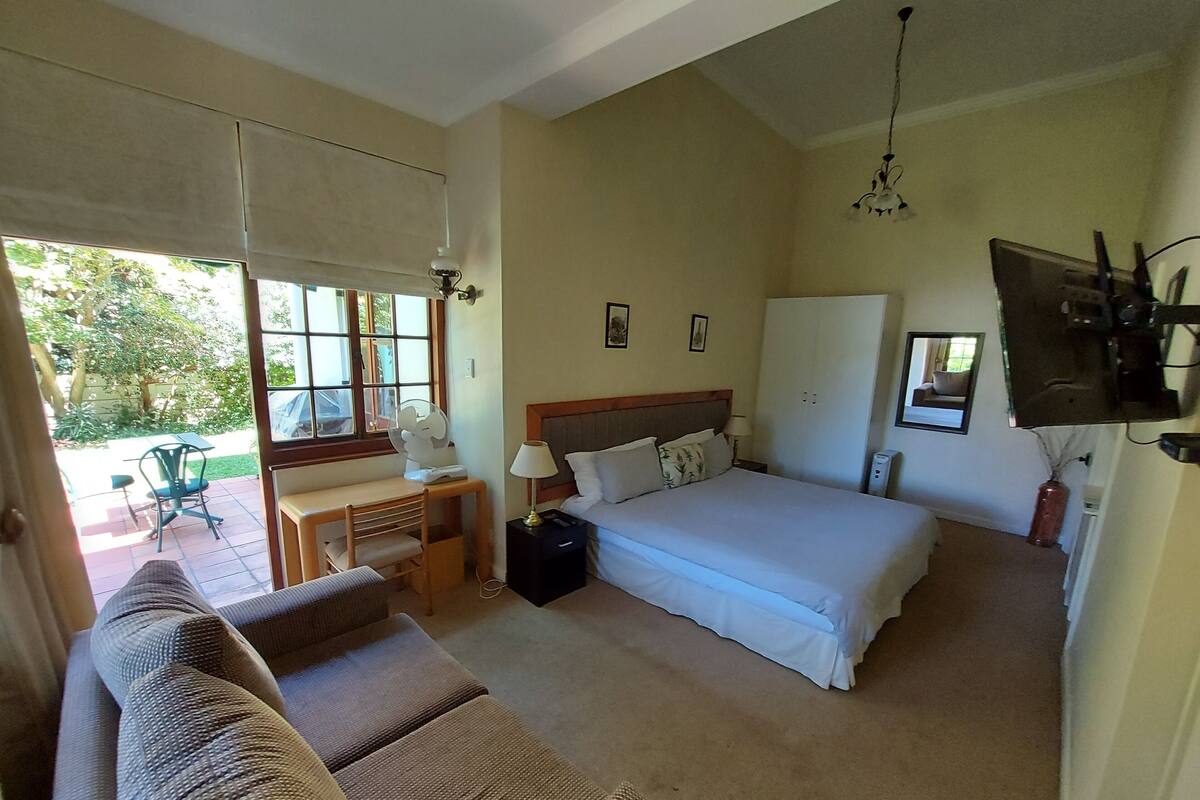 A bedroom with a bed, bedside table, lamp, and a TV. There is a window with blinds, a chair, and a table outside.