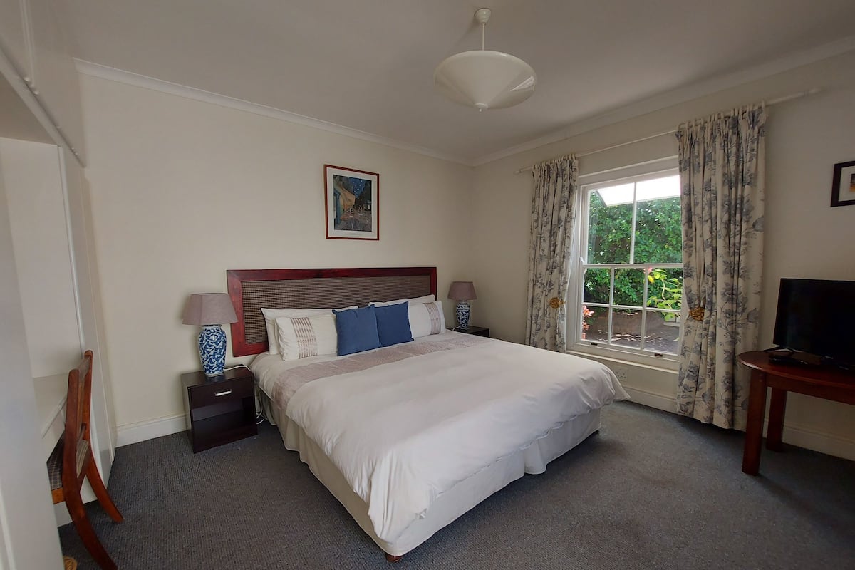 A neatly arranged bedroom with a large bed, a bedside table with lamps, a television on a side table, and a window with curtains.