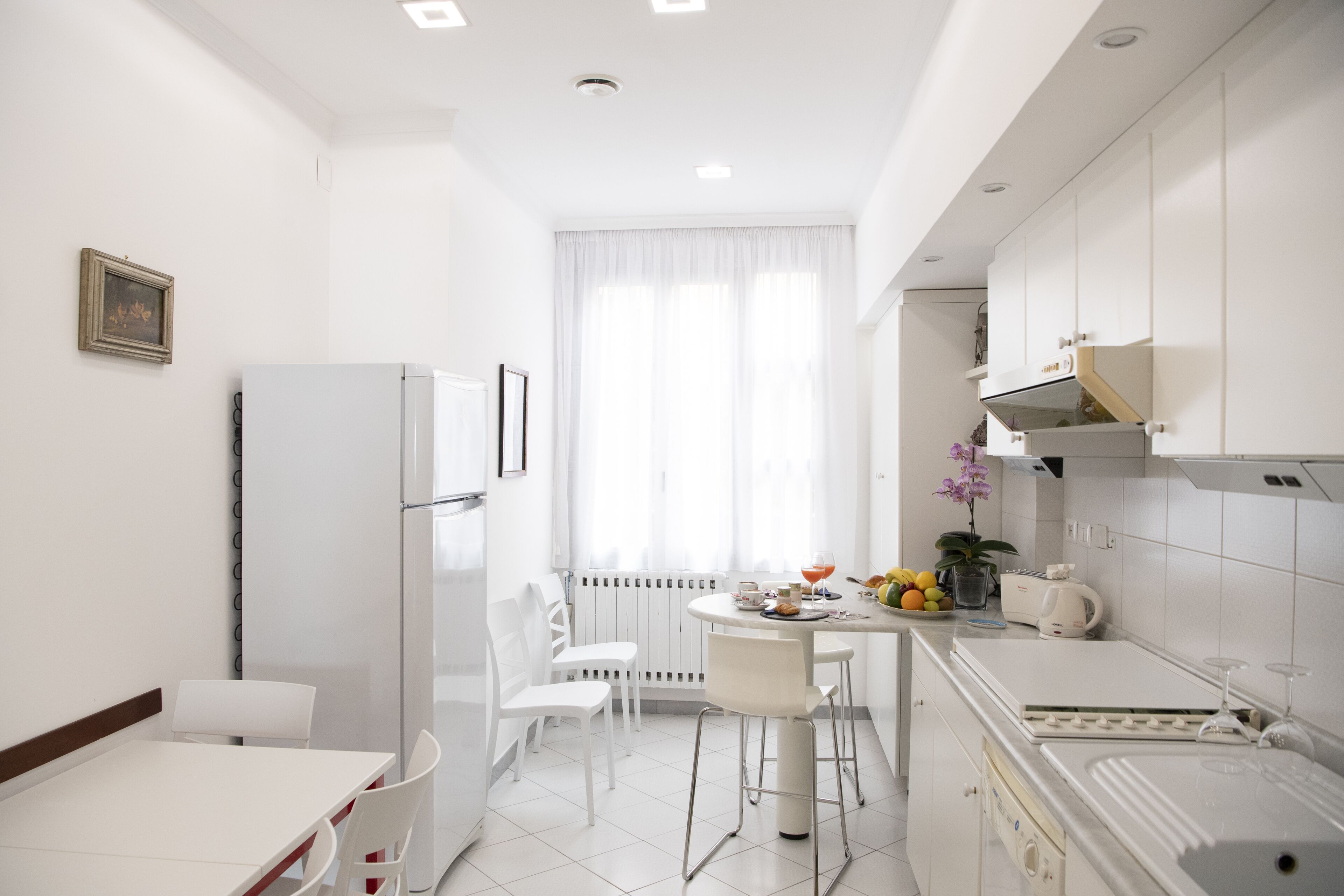 Cozinha moderna com armários brancos, área de jantar com mesa e cadeiras brancas, e janela com cortinas leves.
