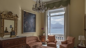 Une chambre avec un grand lit, deux fauteuils, une commode, un grand tableau, un lustre, une fenêtre donnant sur la mer et une petite table avec une lampe.