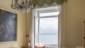 Une chambre à coucher avec un grand lit, deux fauteuils, un lustre, un tableau et une vue sur la mer.