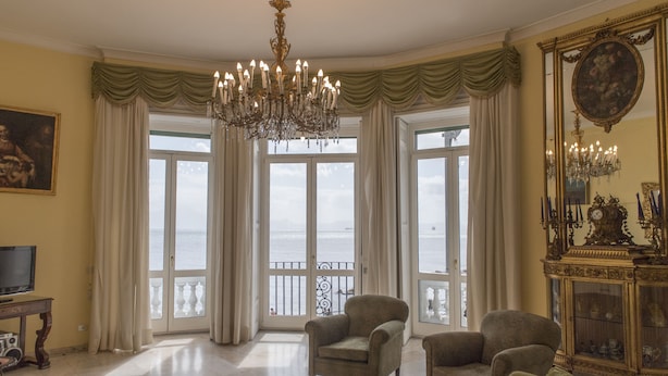 Un vaste salon agrémenté d’un grand lustre, de deux fauteuils, d’un téléviseur et offrant une vue sur la mer grâce à de grandes fenêtres.