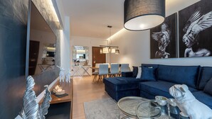 Una sala de estar moderna con un sofá azul, una mesa de centro de vidrio y una zona de comedor con una mesa y sillas de madera.