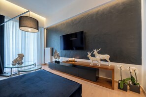 A modern living room with a flat-screen TV mounted on a textured wall, a glass coffee table with a pug figurine, a wooden bench with decorative items, and potted orchids.