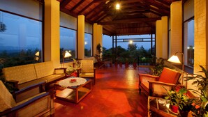 Un amplio patio cubierto con mobiliario de madera, plantas en macetas y ventanas grandes que ofrecen vistas al exterior.