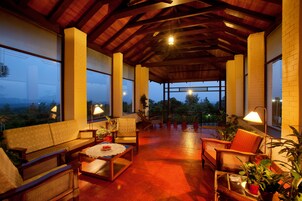 Un amplio patio cubierto con mobiliario de madera, plantas en macetas y ventanas grandes que ofrecen vistas al exterior.