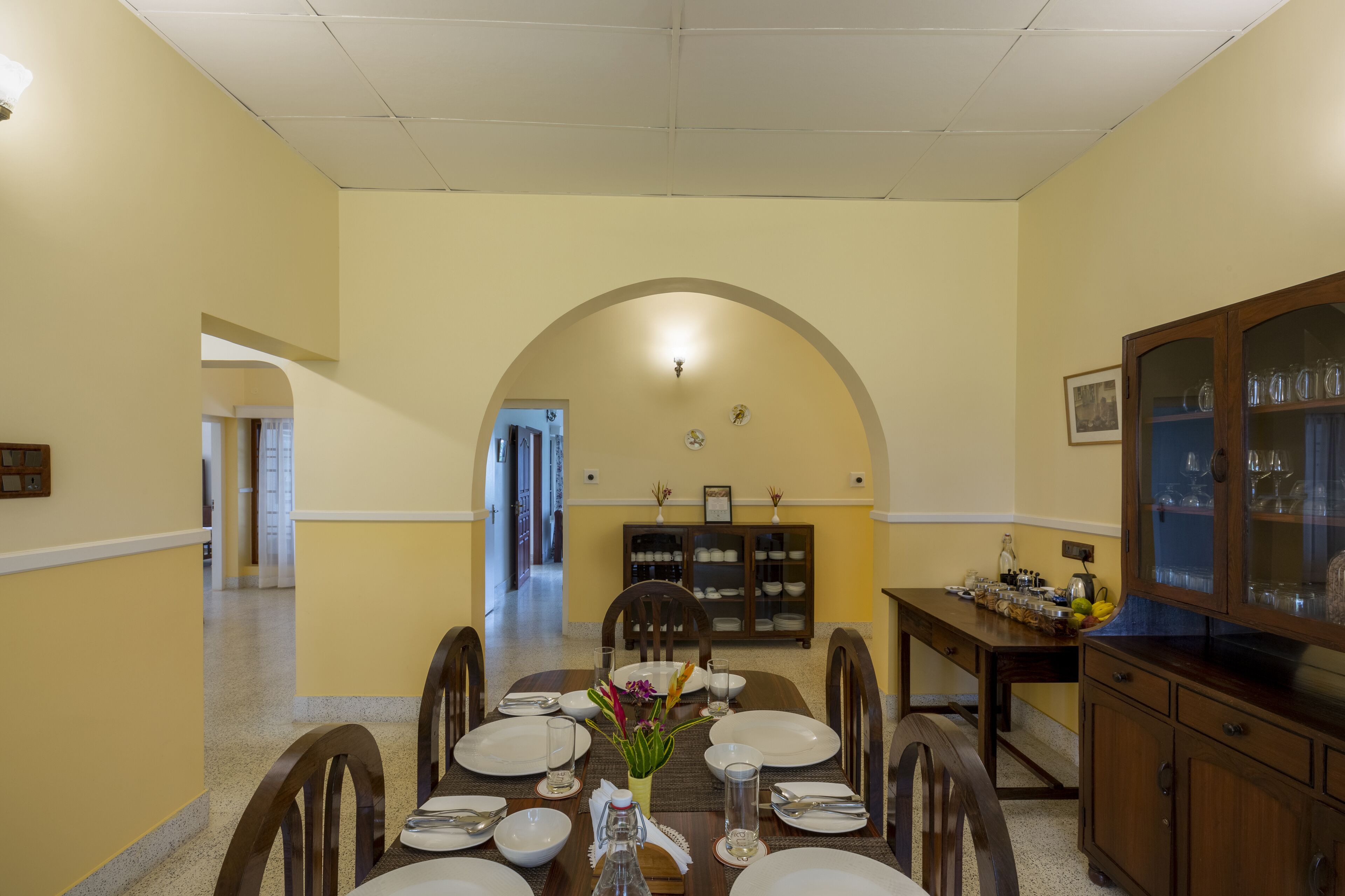 Una sala da pranzo con un lungo tavolo apparecchiato per sei persone, circondato da sedie in legno. Sulla destra si trova un mobile con ante in vetro, e una porta conduce in un'altra stanza.