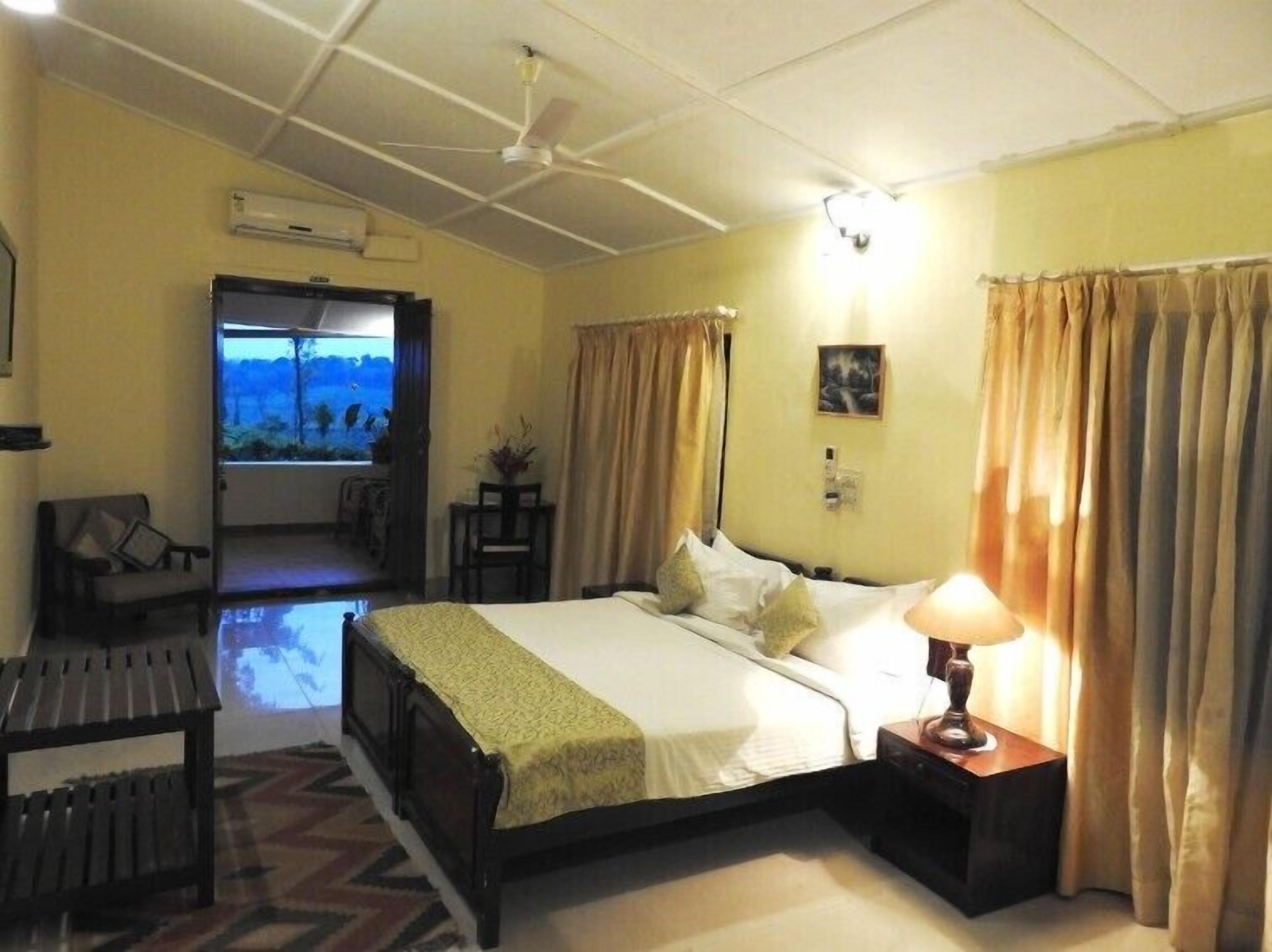 Una habitación de hotel con cama, lámpara de noche, ventana con cortinas, una silla y vistas al exterior.