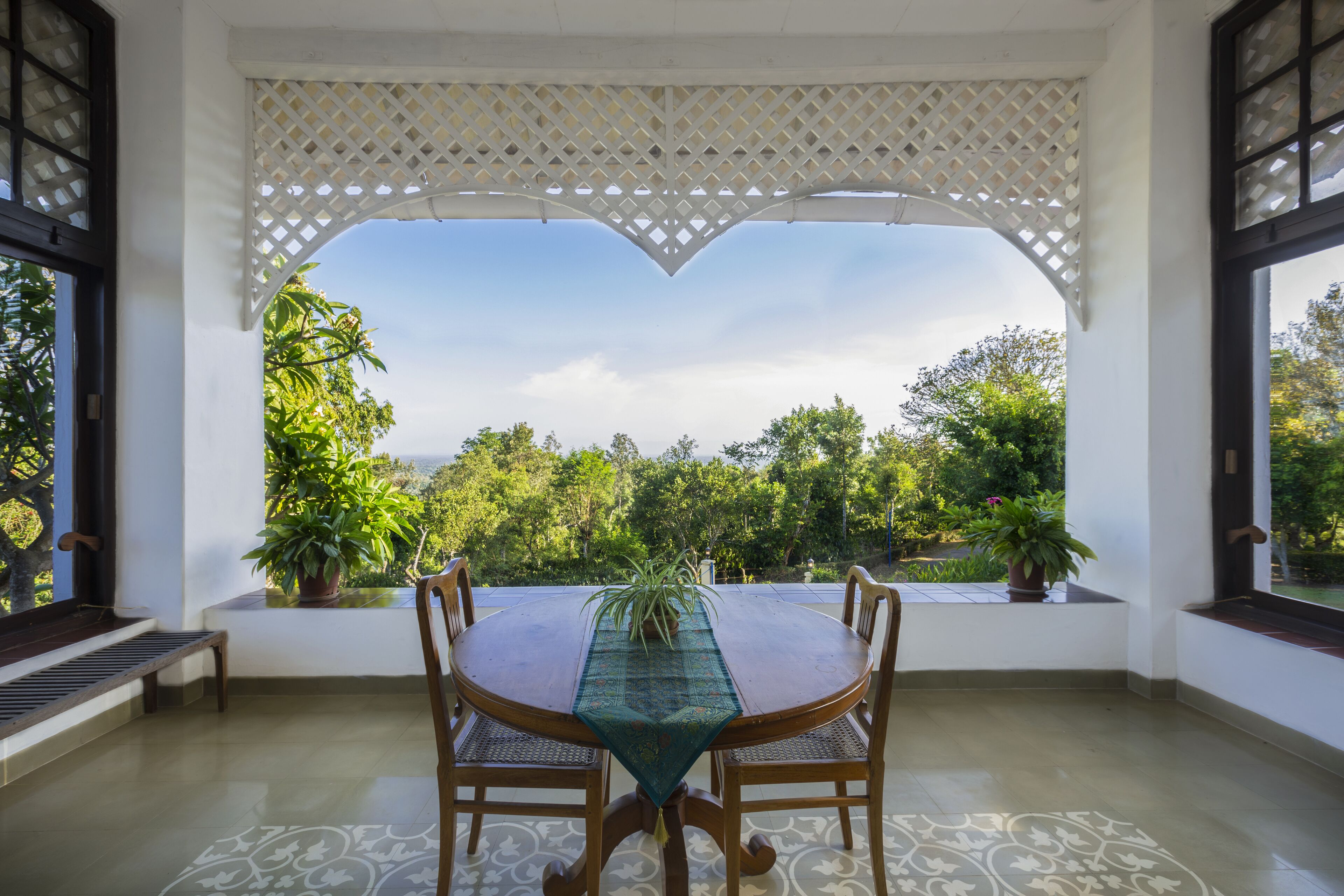 Uma área de refeições com mesa e cadeiras, vista para o verde através de uma janela arqueada e piso estampado.