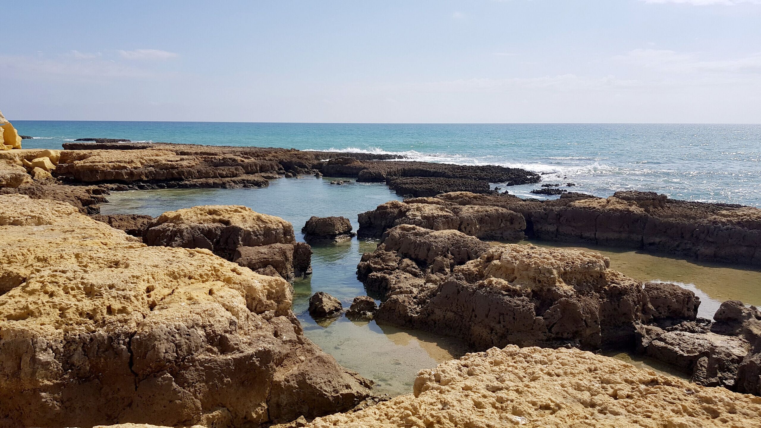 A rocky coastal landscape with clear blue water and a sandy foreground.