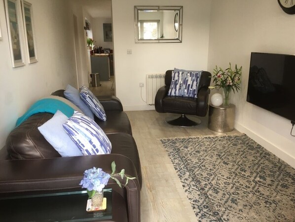A living room with a dark brown leather sofa, a black armchair, a large flat-screen TV, a patterned rug, and a small vase with flowers. -   LUXURY 1 BED  FLAT  WITH GARDEN & PARKING - 2 MINS WALK TO RIVER  DOG FRIENDLY (Bournemouth)