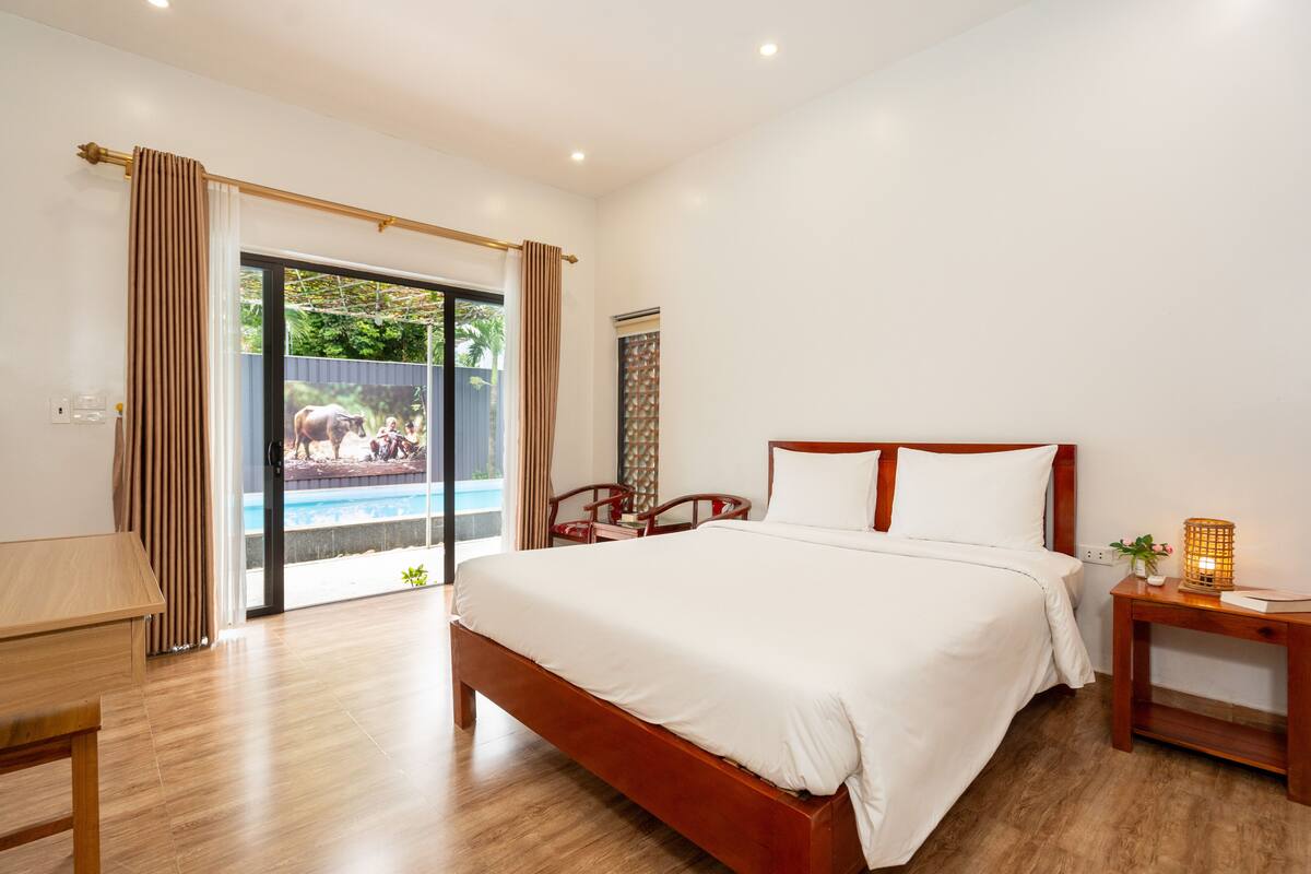 A bedroom with a large bed, a wooden desk, a nightstand, and a view of an outdoor area with a pool and a cow.