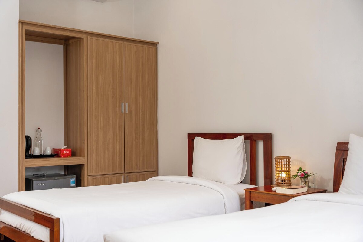 Habitación de hotel con dos camas individuales, un armario de madera, una mesita de noche con lámpara y un pequeño estante con artículos de aseo.