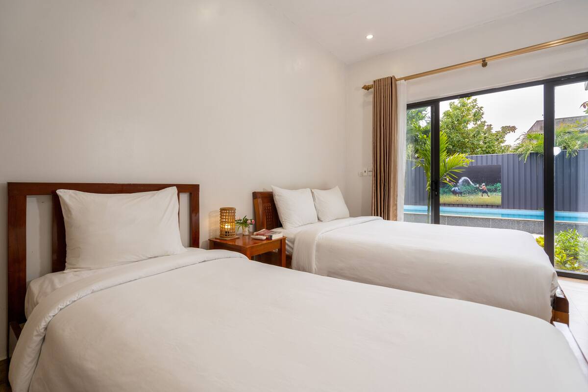 A hotel room with two beds, a nightstand with a lamp, and a large sliding glass door leading to a pool area.