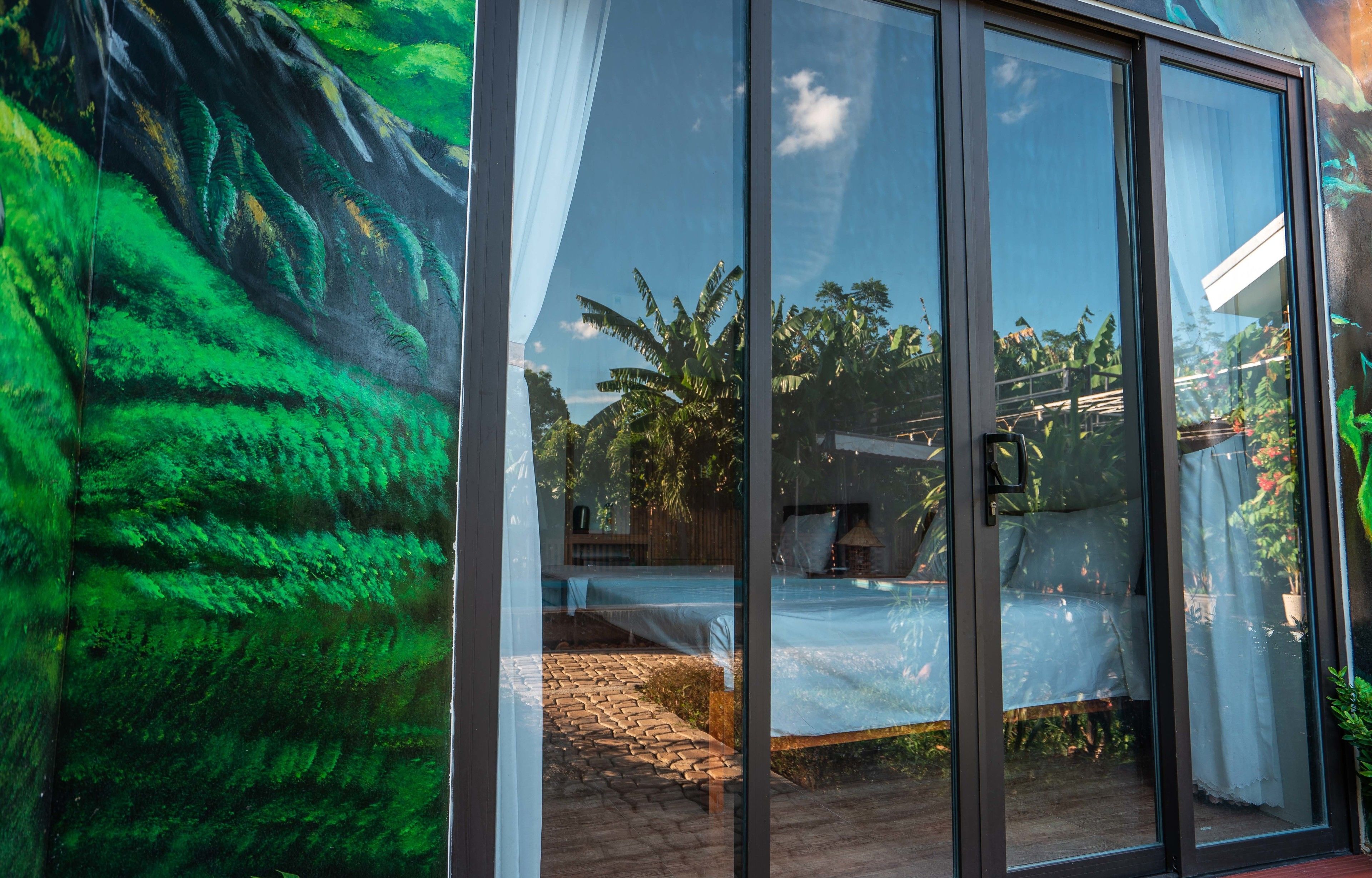 A modern room with large glass doors, a bed with a white sheet, and a view of lush greenery outside.