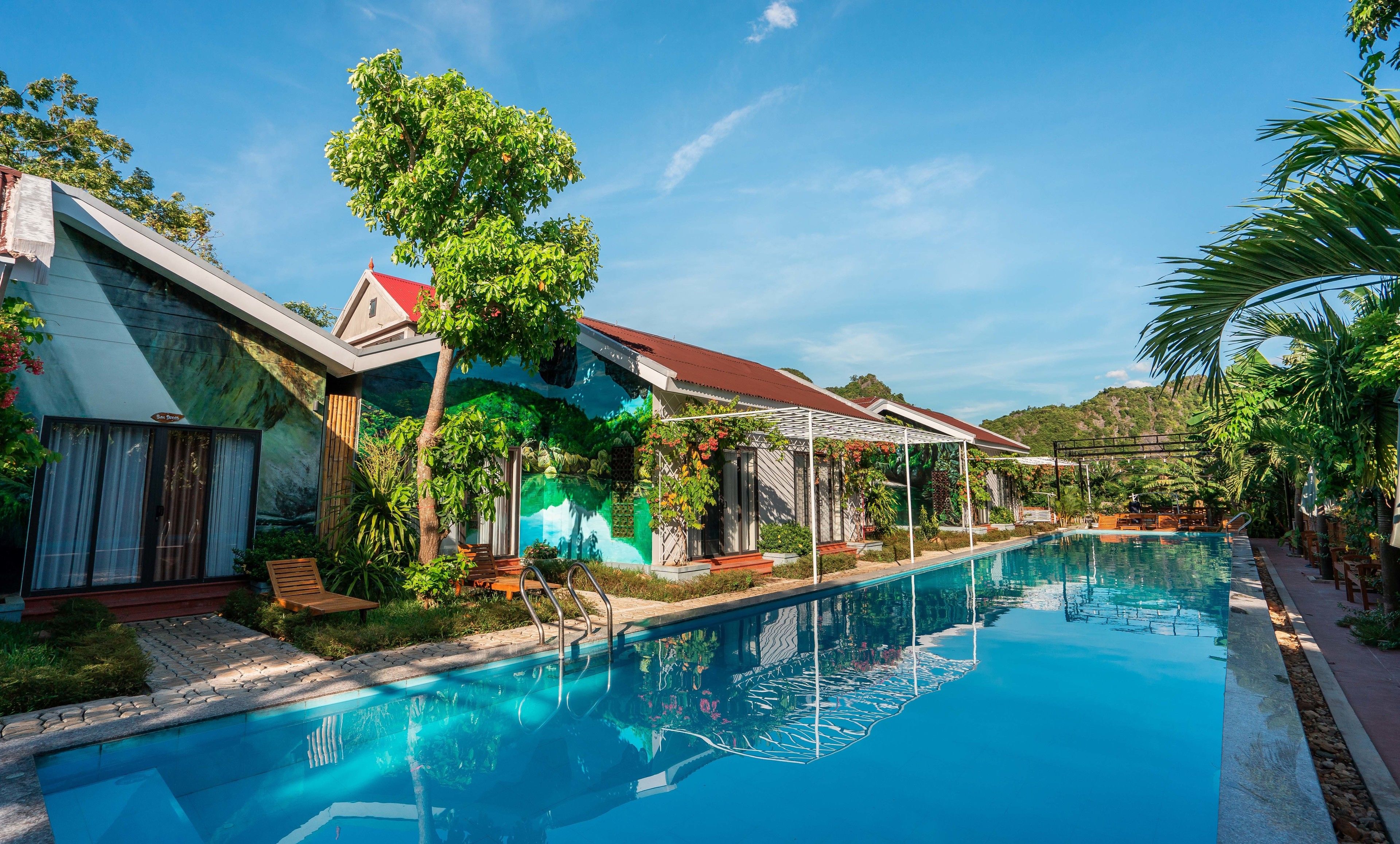 A swimming pool with a clear blue water surface, surrounded by lush greenery and a building with a red roof.