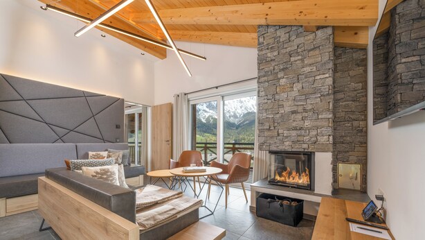 Un salon moderne doté d’une cheminée en pierre, de meubles en bois et offrant une vue sur les montagnes grâce à de grandes fenêtres.