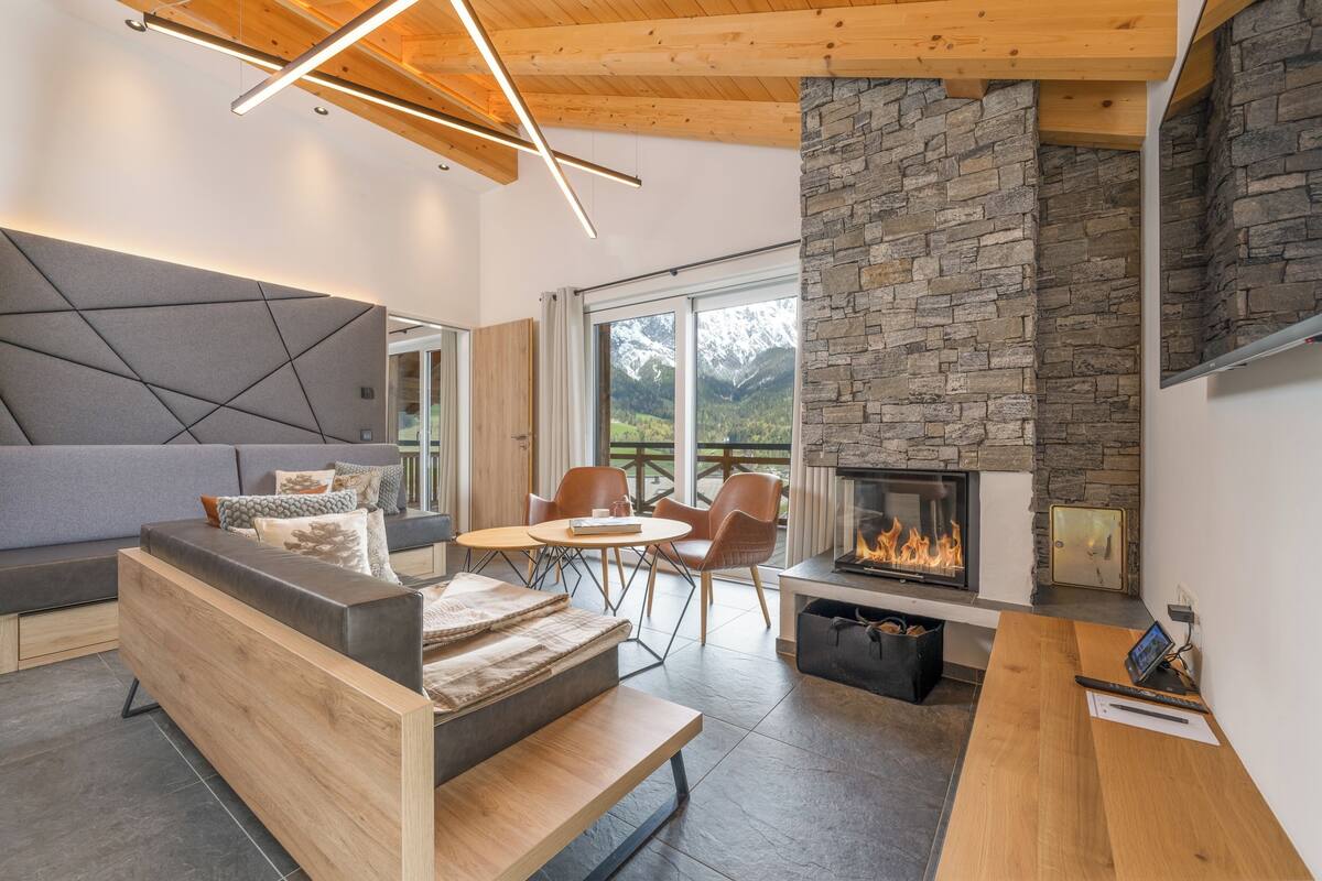 Ein modernes Wohnzimmer mit Kamin aus Stein, Holzmöbeln und Blick auf Berge durch große Fenster.
