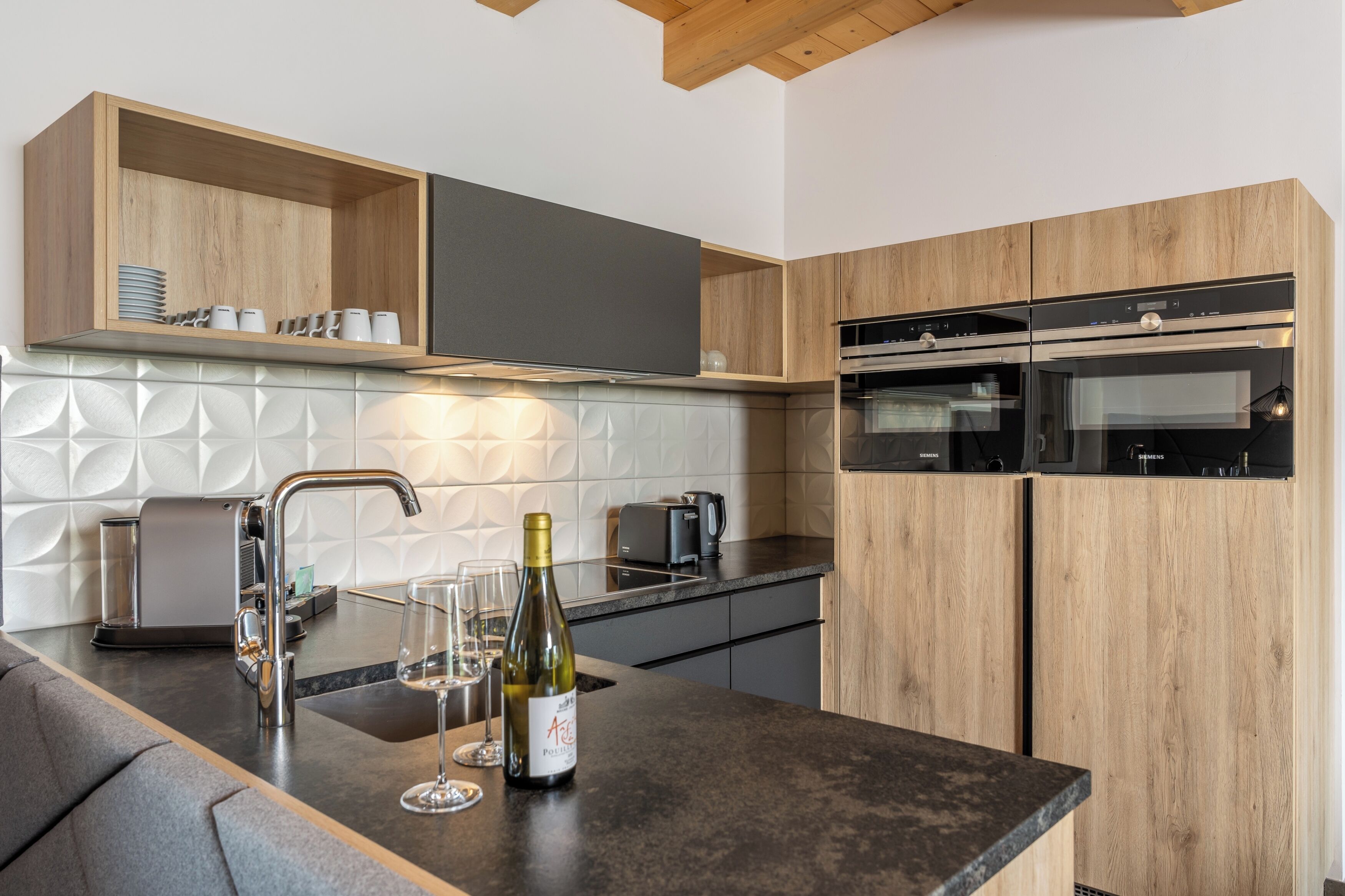A modern kitchen with wooden cabinets, a built-in oven, a wine bottle, and glasses on the counter.
