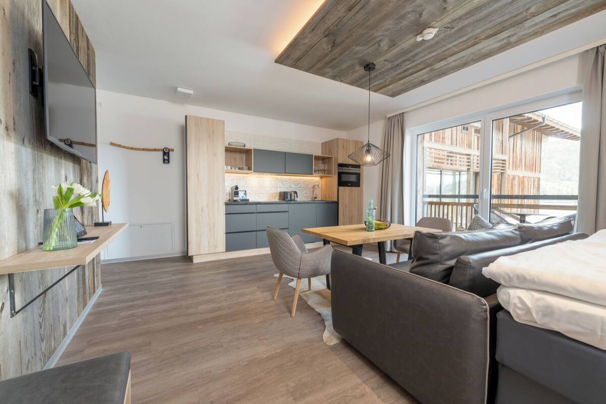 Eine moderne Küche mit Holzschränken, ein Essbereich mit Tisch und Stühlen sowie eine Sitzecke mit Sofa und großem Fenster.