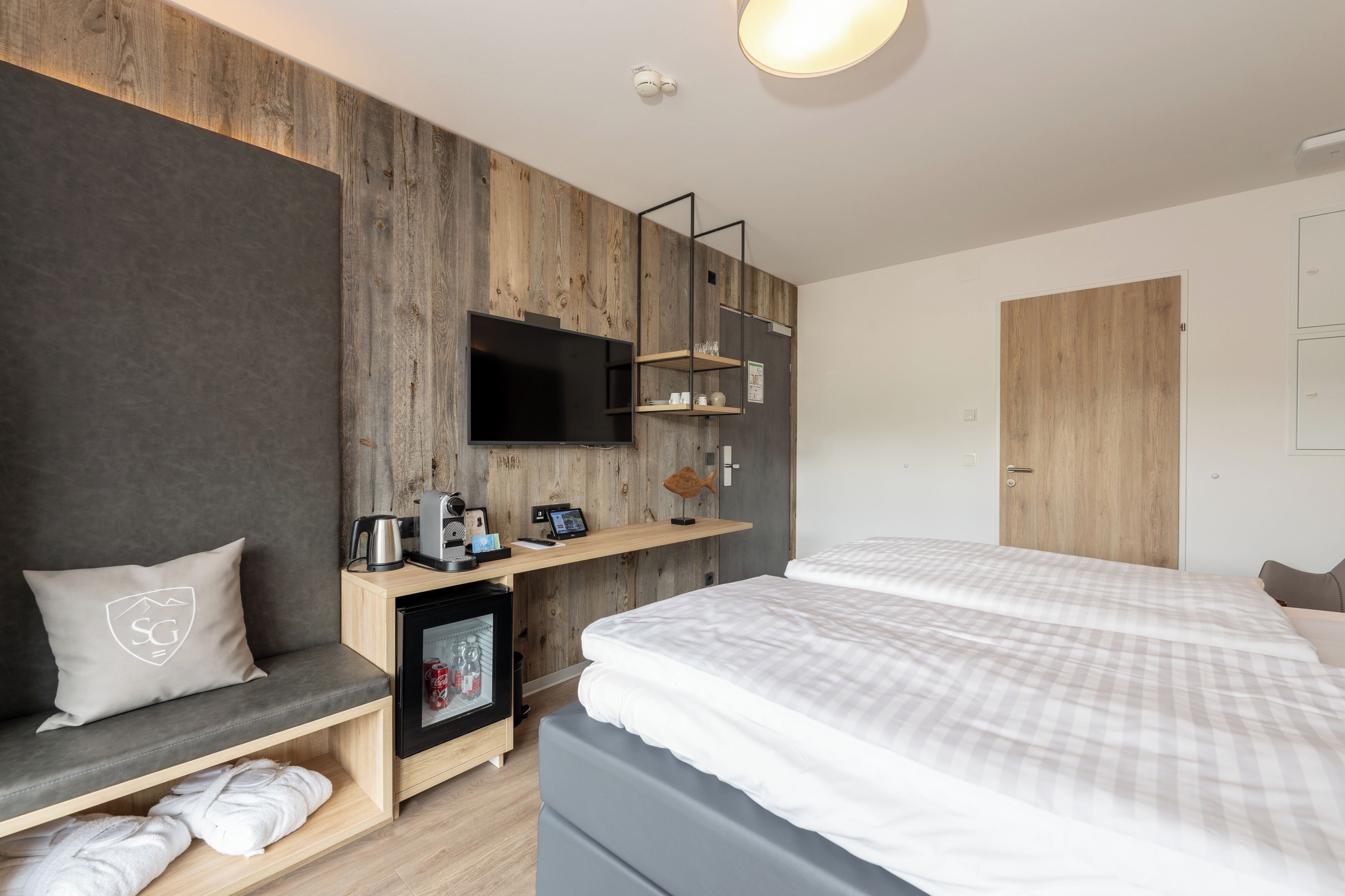 Ett modernt hotellrum med en väggpanel i trä, en platt-TV, ett skrivbord med en kaffebryggare, en inramad tavla och en säng med vita sängkläder.