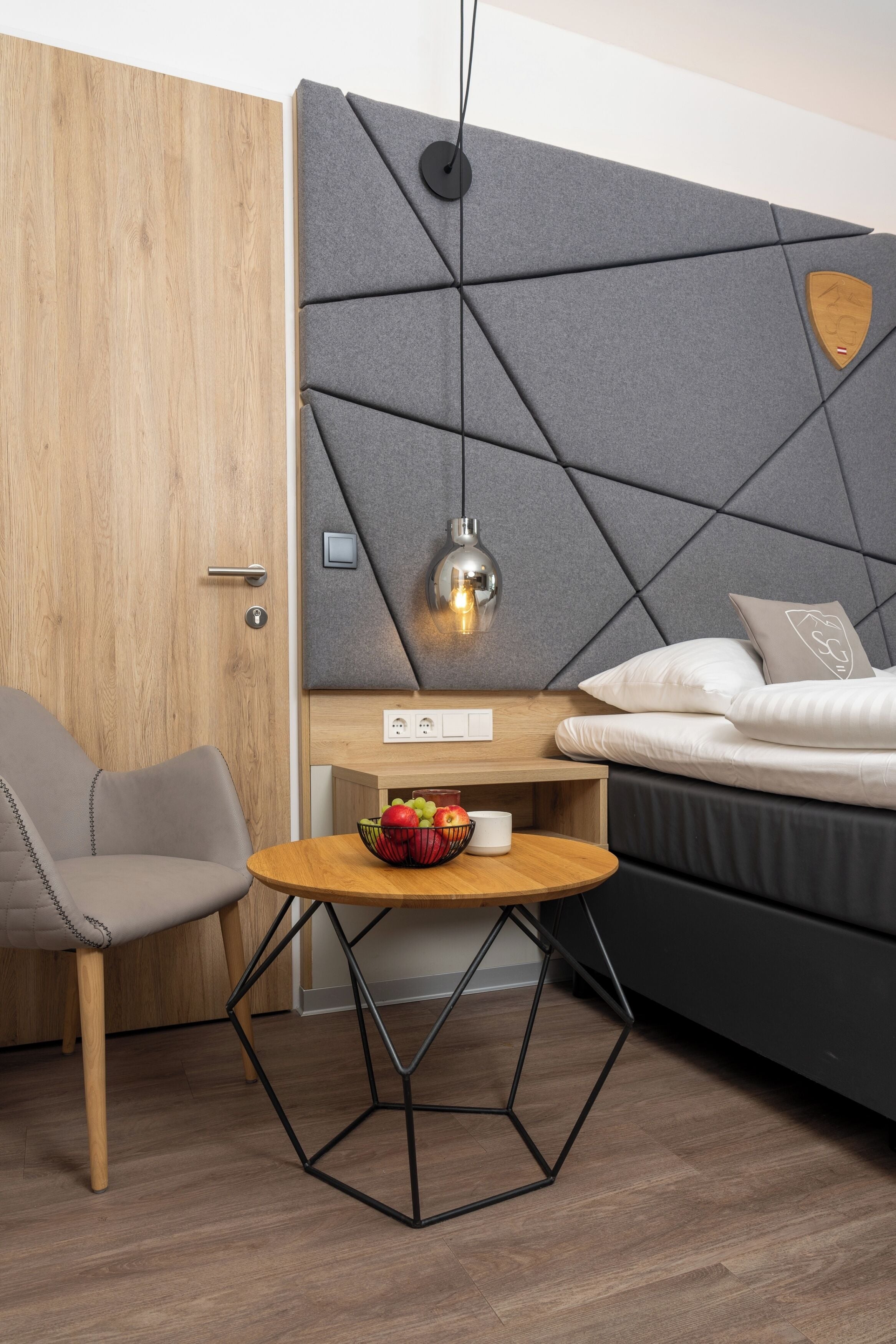 A modern bedroom with a wooden door, a grey headboard with a geometric pattern, a bed with white bedding, a small round table with a bowl of fruit, and a hanging pendant light.