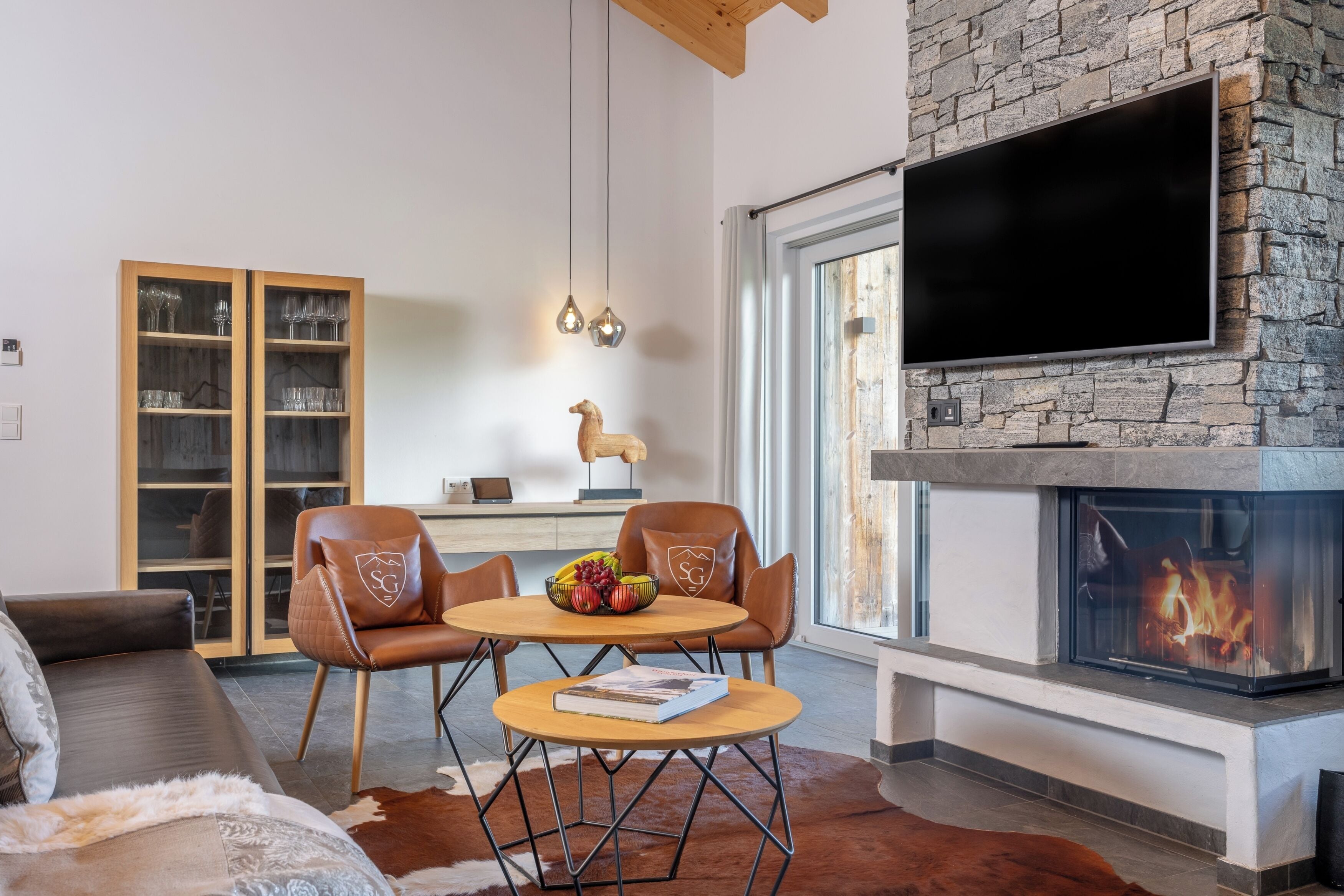 A modern living room with a fireplace, a television mounted on a stone wall, a wooden cabinet with glass doors, and a cozy seating area with leather chairs and a round table.