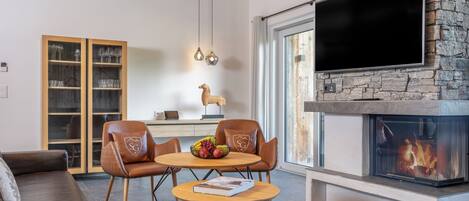 A modern living room with a fireplace, a television mounted on a stone wall, a wooden cabinet with glass doors, and a cozy seating area with leather chairs and a round table.