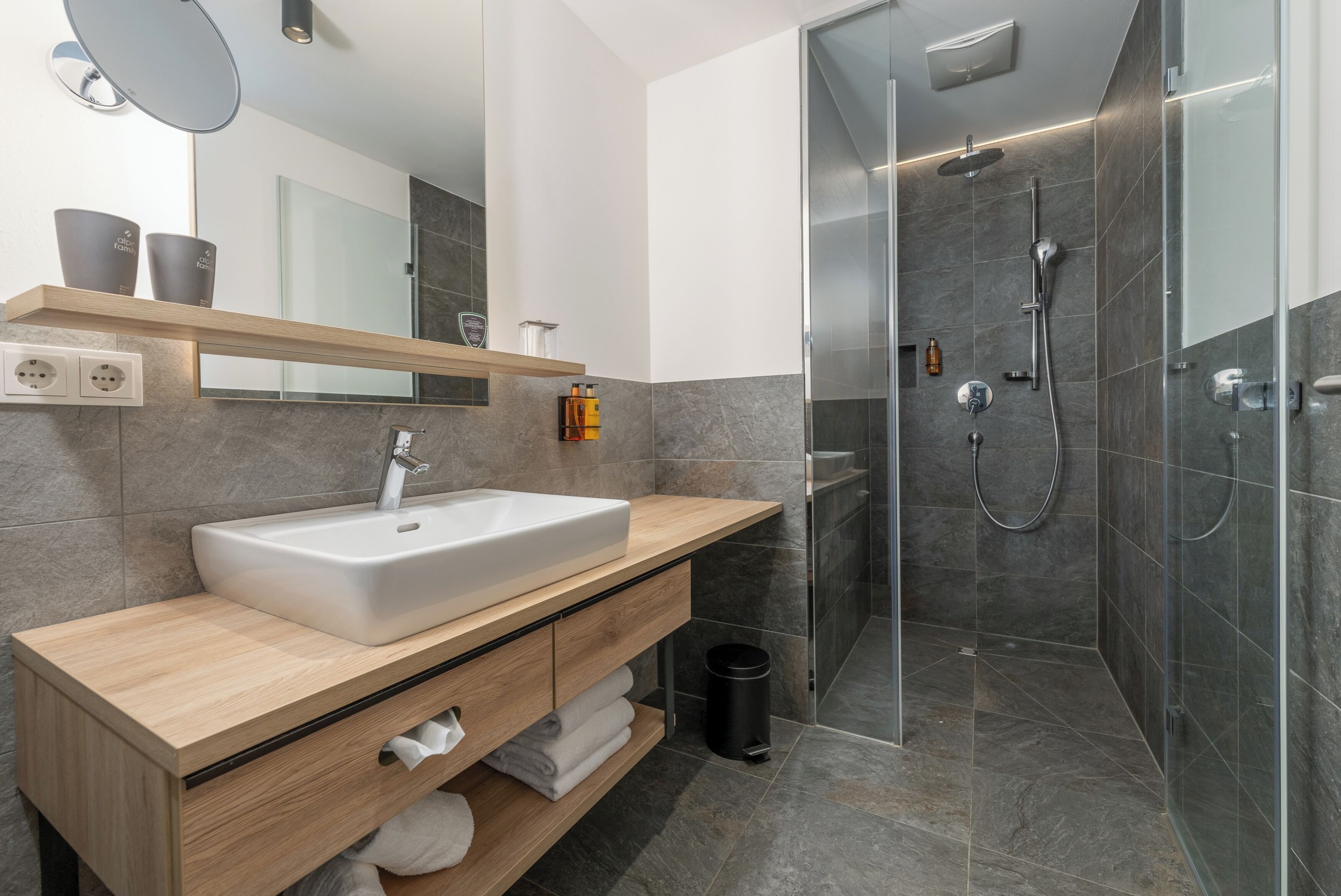 A modern bathroom with a wooden vanity, a white sink, a mirror, and a glass-enclosed shower area.