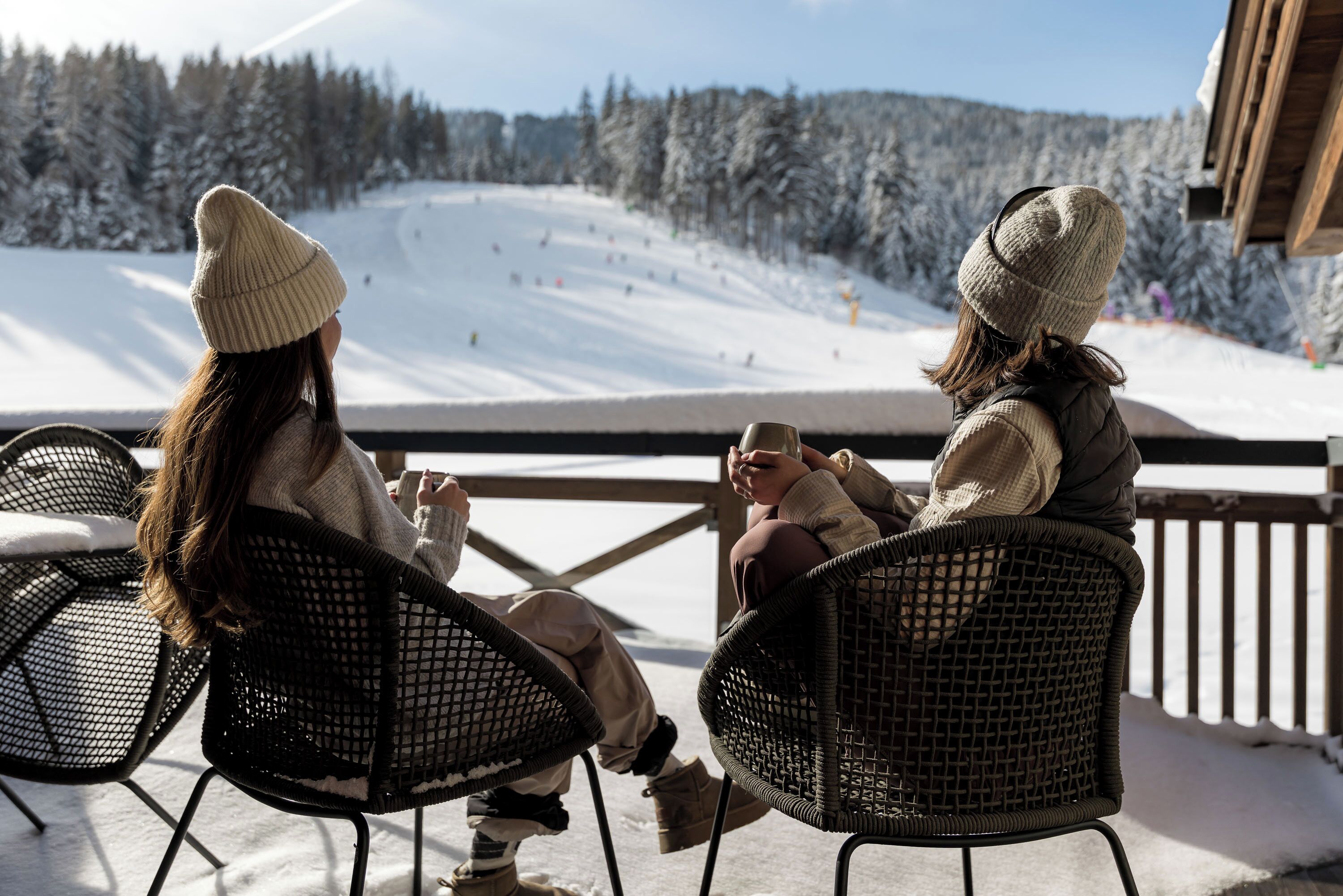 Two people sitting on outdoor chairs facing a snowy ski slope.