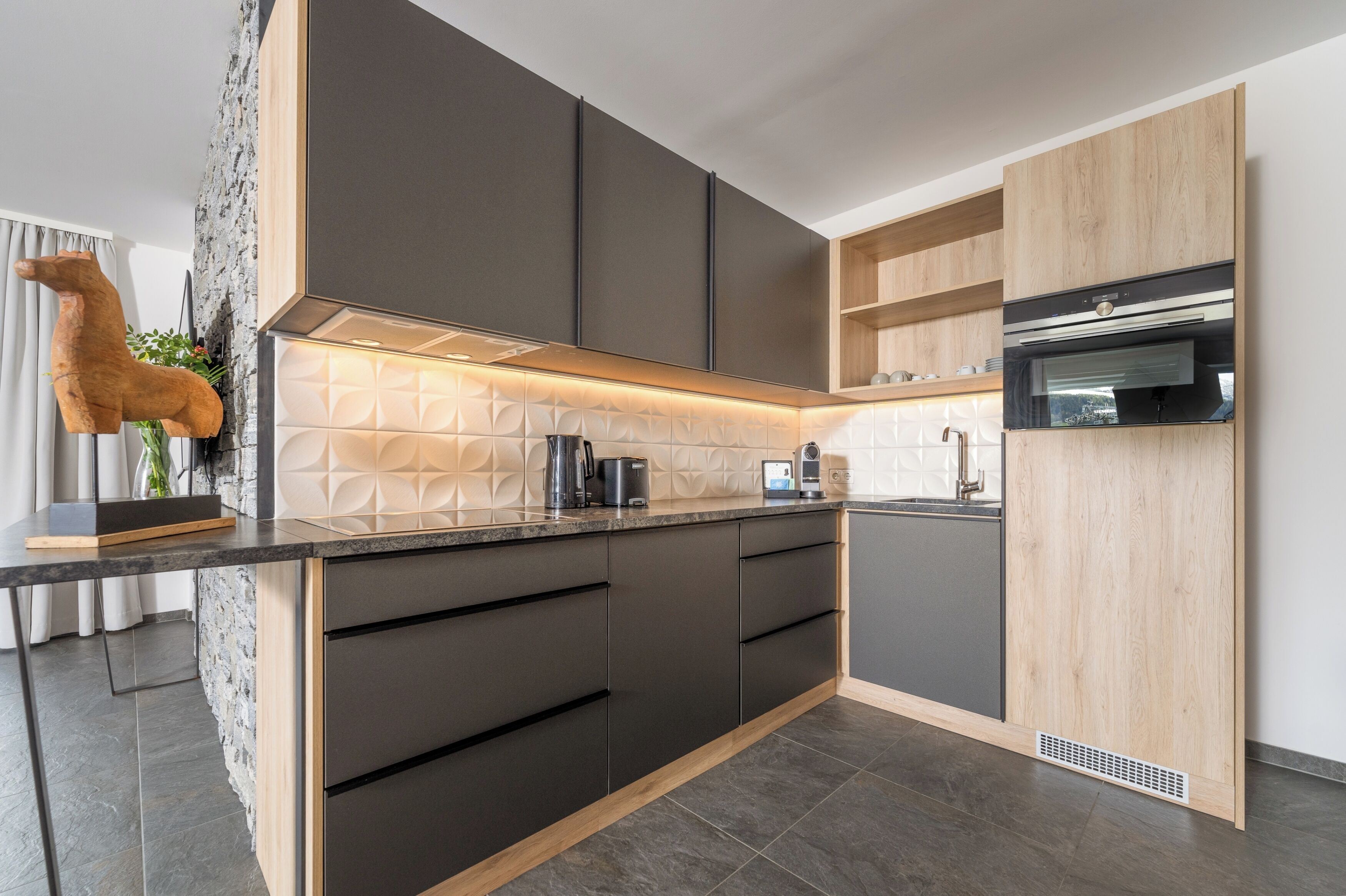 Una cocina moderna con armarios oscuros, horno empotrado y salpicadero de piedra.