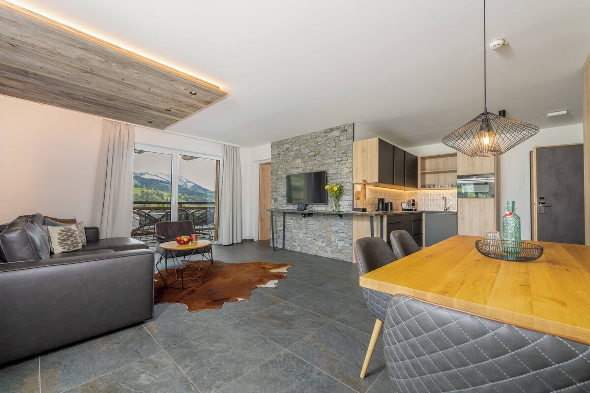 Ein modernes Wohnzimmer mit einer Couch, Esstisch und Stühlen. Es gibt eine Küchenzeile mit einer Steinrückwand und einen Fernseher. Das Zimmer hat ein großes Fenster mit Bergblick.