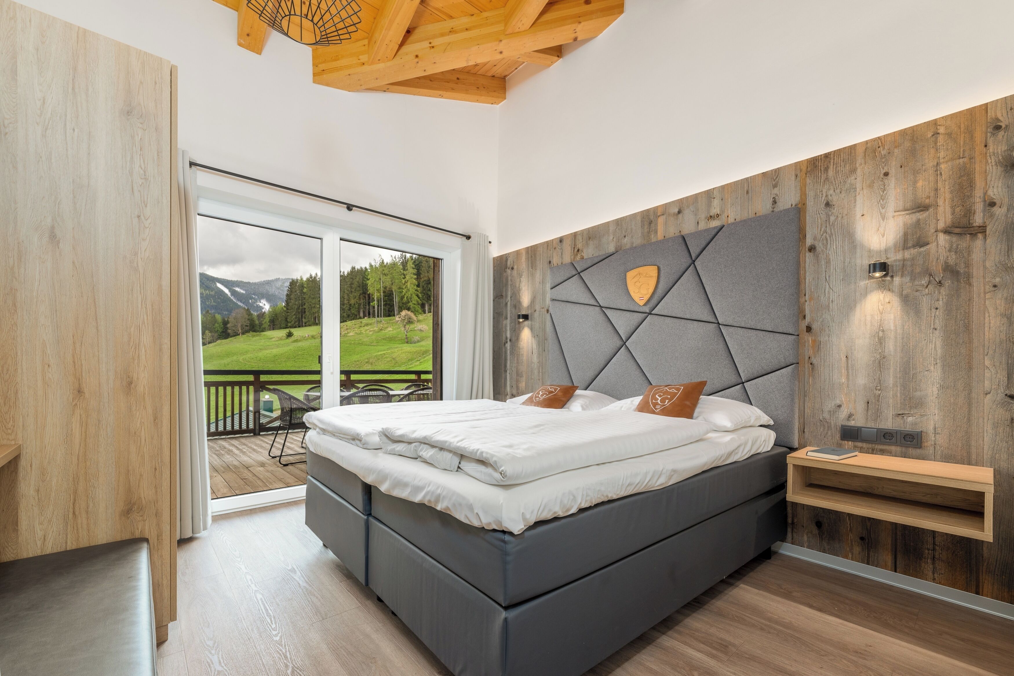 A bedroom with a bed, a wooden headboard, a nightstand, and a view of a balcony with a mountain landscape.