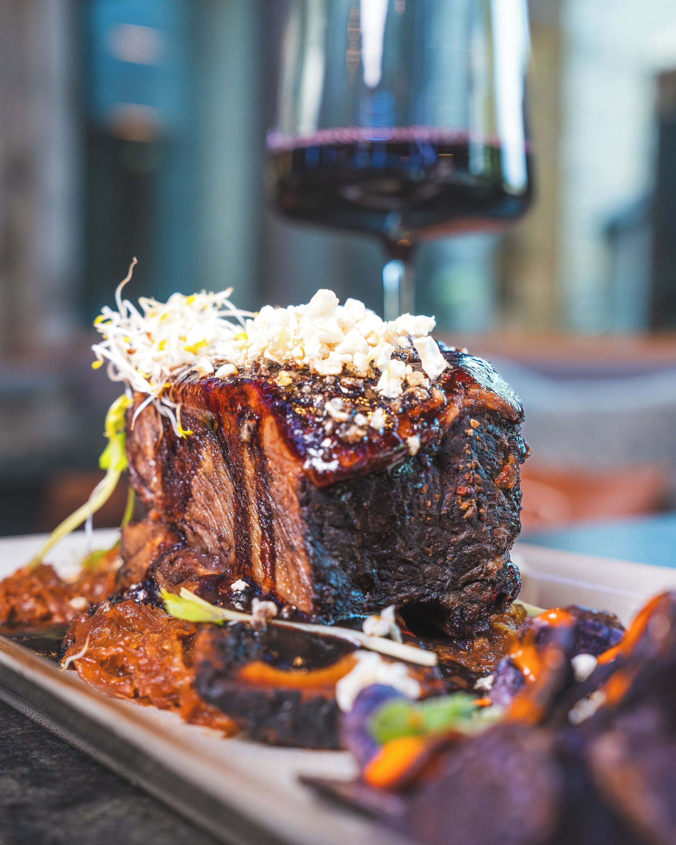 A steak with sprouts and a sauce on a plate, with a glass of red wine in the background.