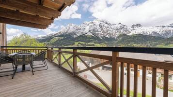Una terraza de madera con una mesa y sillas, con vistas a un paisaje montañoso con picos nevados y cielo despejado.