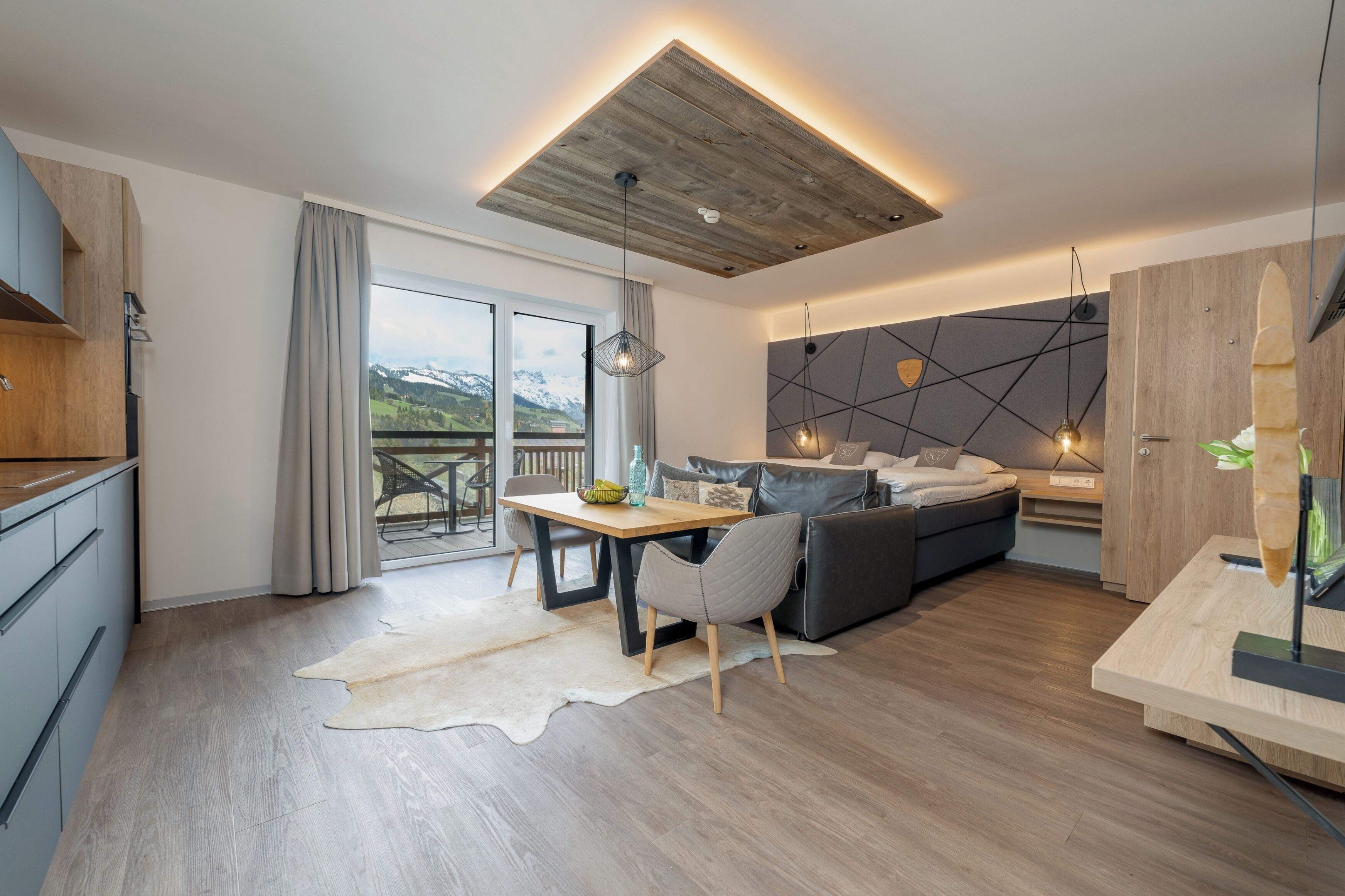 Una sala de estar moderna con techo de madera, un sofá, una mesa de comedor con sillas y vistas a montañas a través de un ventanal grande.